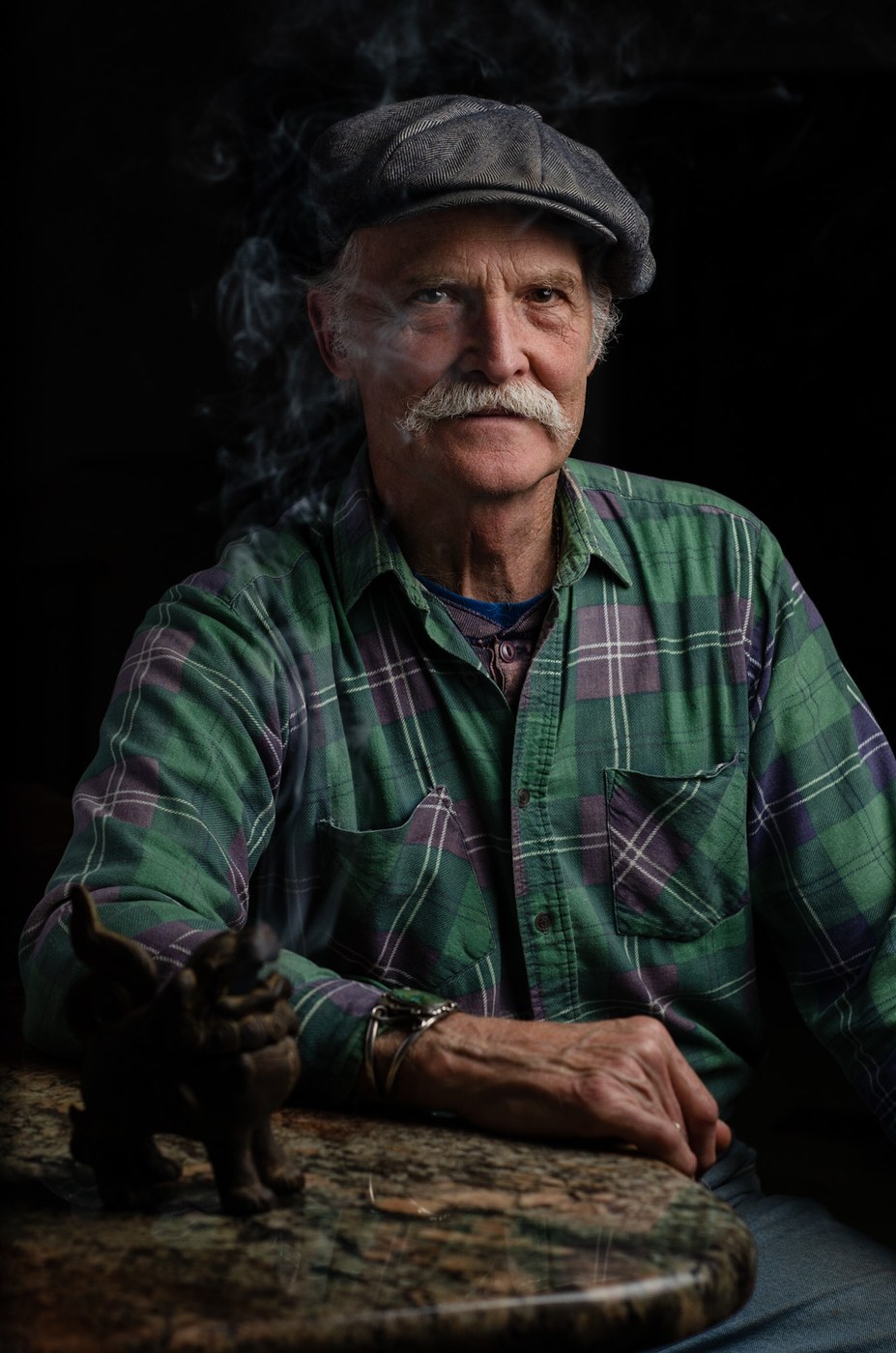 Nathaniel Miller at a granite table in a flat cap and green plaid shirt, smoke curling around him, a small carved figure beside his hand.