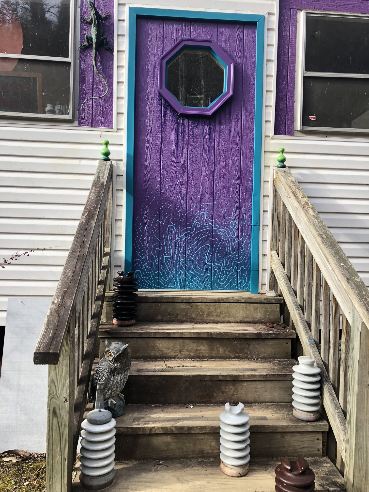 A purple side door with a teal frame and an octagonal window, blue swirls painted at its base, at the top of wooden steps.