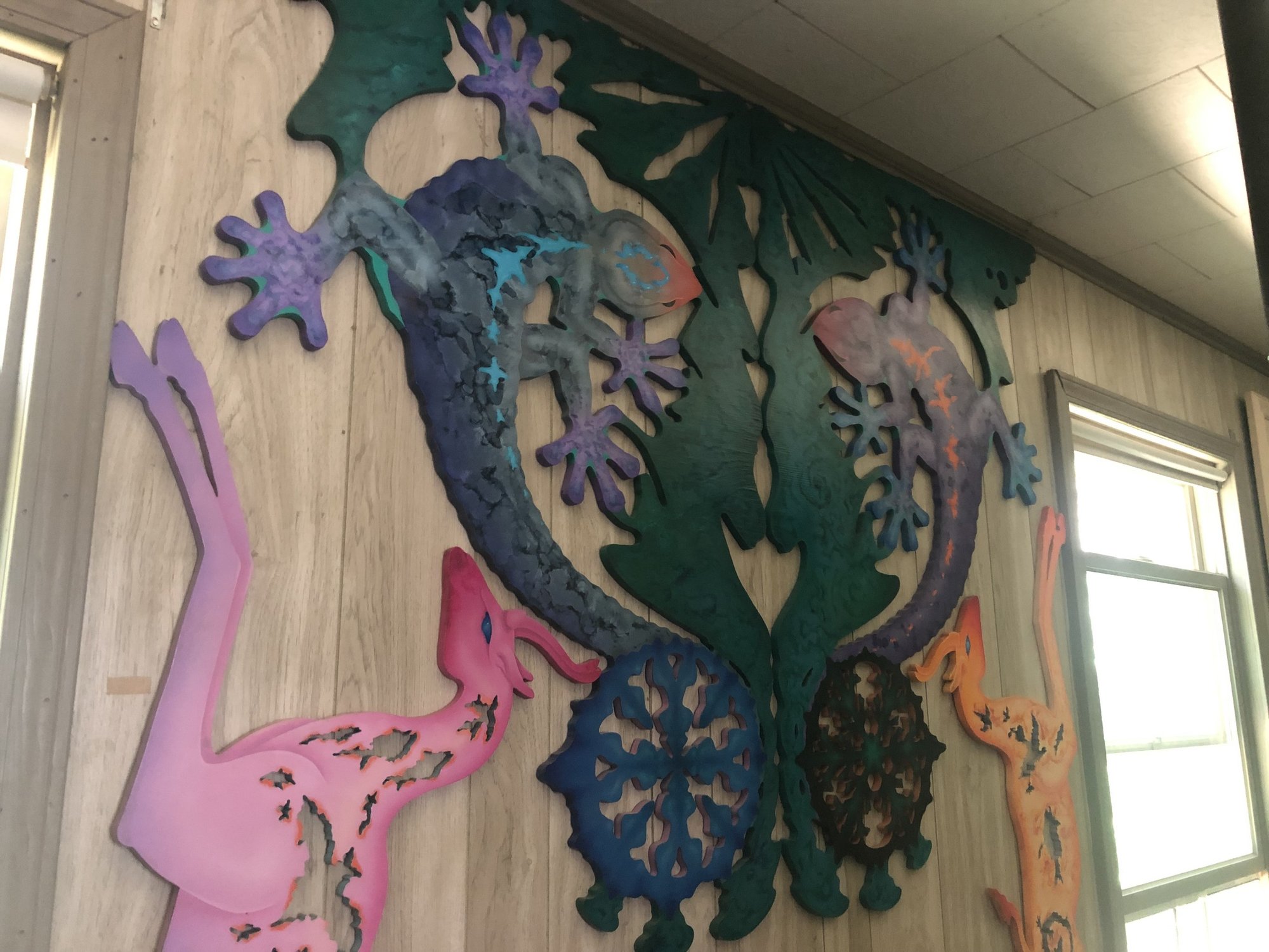 Two carved and painted geckos face each other on a green leaf cutout, flanked by a pink and an orange leaping animal, on a wood-paneled wall between two windows.