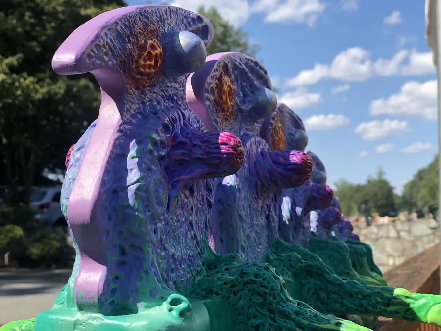 A row of purple, lavender and teal carved forms with glowing orange-centered blossoms, in strong sunlight against a blue sky.