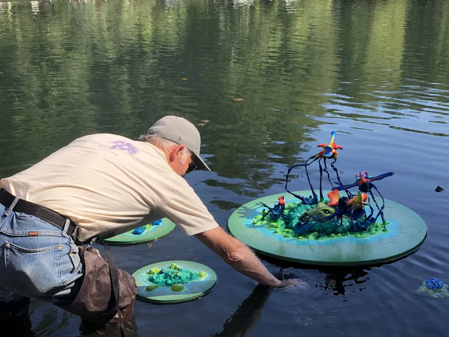 A man in a tan cap and cream T-shirt stands in the pond reaching toward a floating green disc that carries a sculpture of blue and orange creatures, two smaller discs floating nearby.