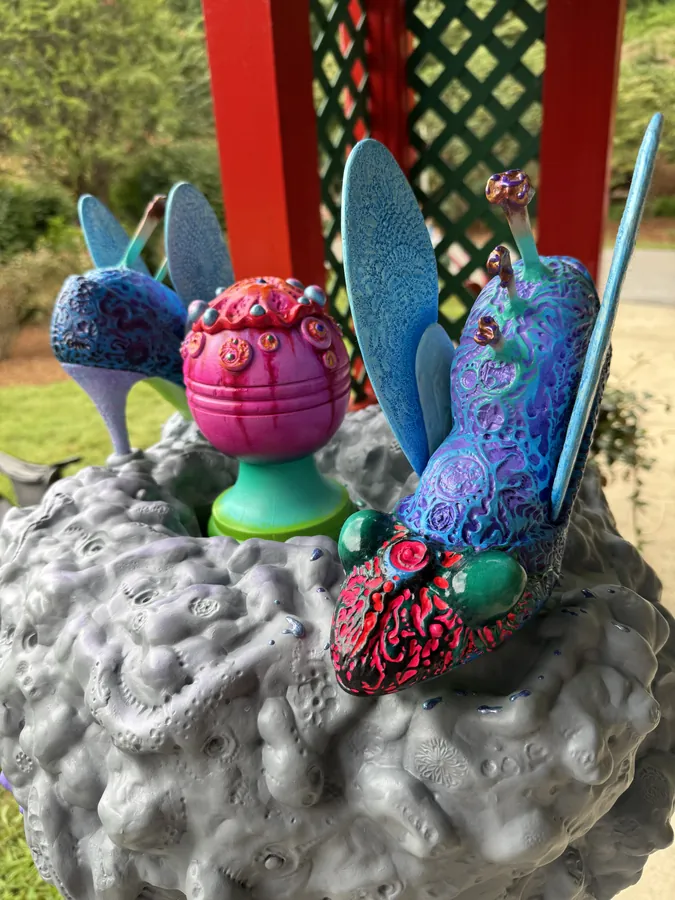 Detail of Big Buzz Part II: two blue winged creatures and a pink orb on the grey, pitted mass at the top.