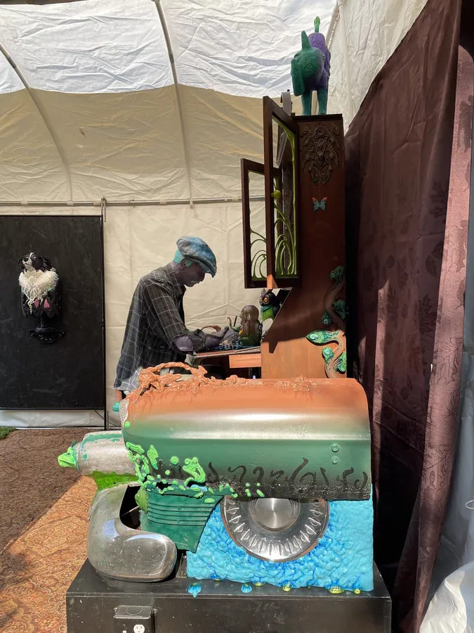Homework for Aunt Louise from the side in the show tent: the figure bends over its desk in the hutch above the car's rounded green, copper and turquoise front, a dog bust on a black wall behind.