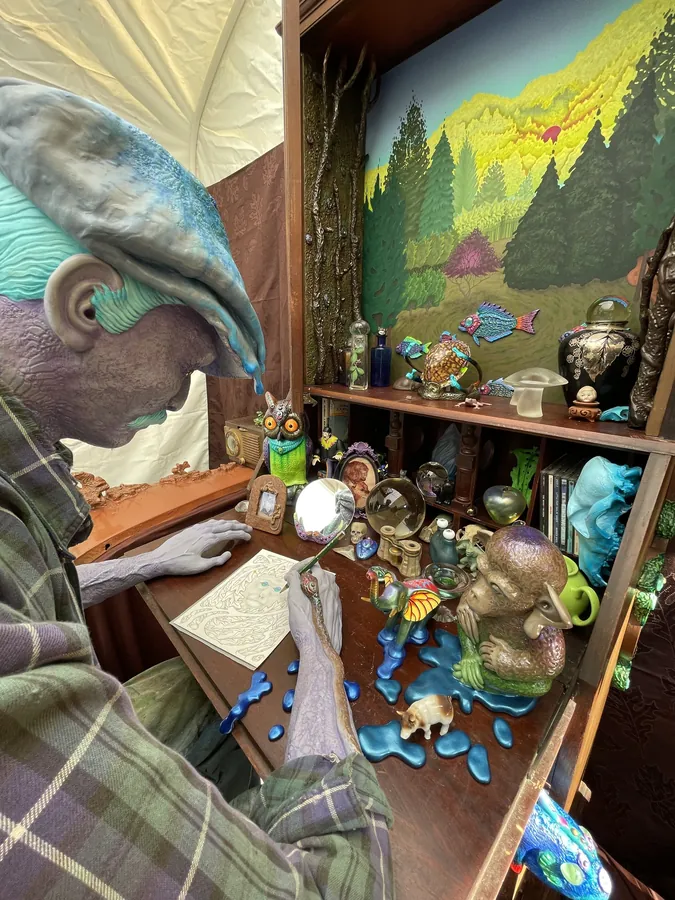 Close view of the seated figure in its flat cap, leaning over a desk crowded with small creatures, a hand mirror and a teapot, a painted landscape on the wall behind.
