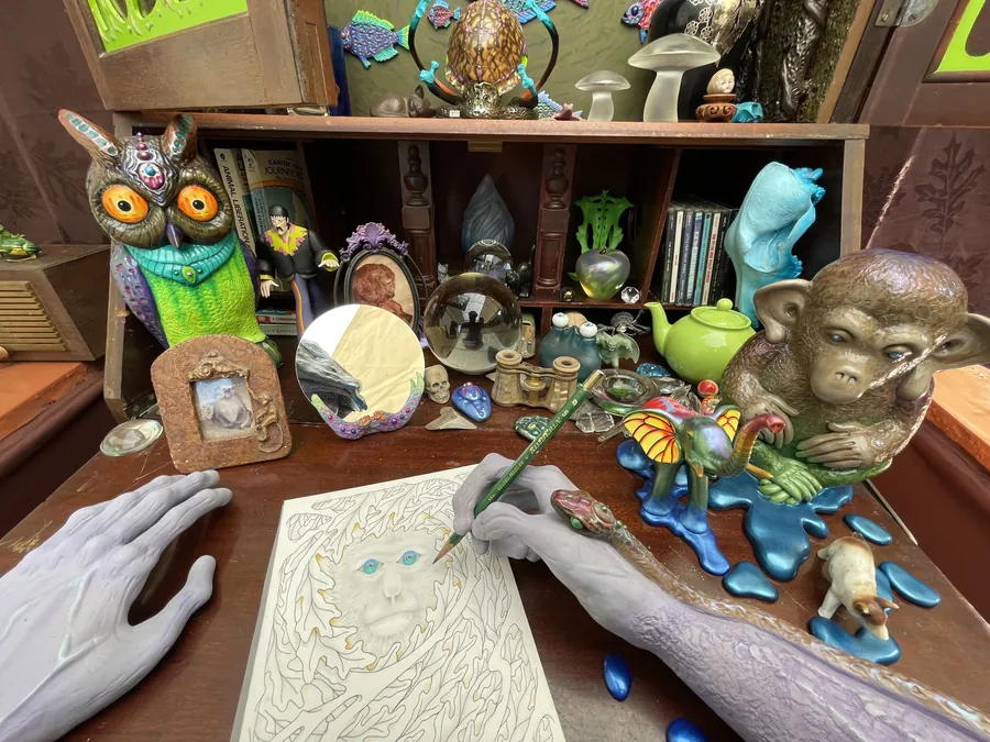 The desk up close: two grey hands, one drawing with a pencil on a sheet of white line art, among an owl, a monkey, a teapot, framed pictures and tiny animals on blue puddles.