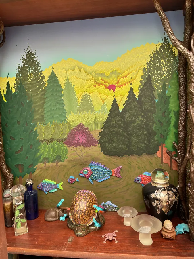 The painted landscape inside the hutch: dark green trees on yellow hills under a red sun, small fish swimming in the meadow, vases and glass mushrooms on the shelf below.