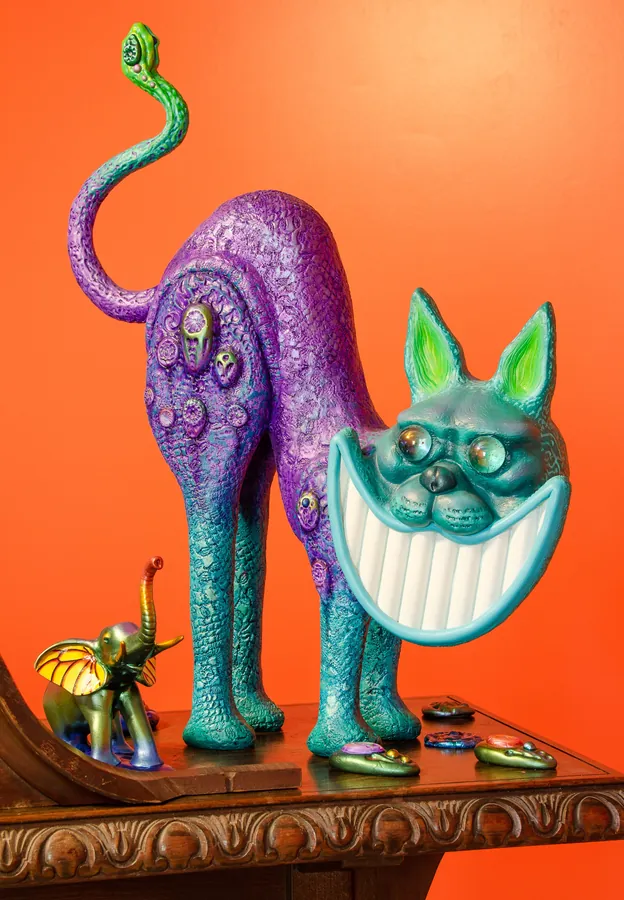 A purple cat with a wide teal grin and a coiled tail arches its back on a carved table, against an orange wall.