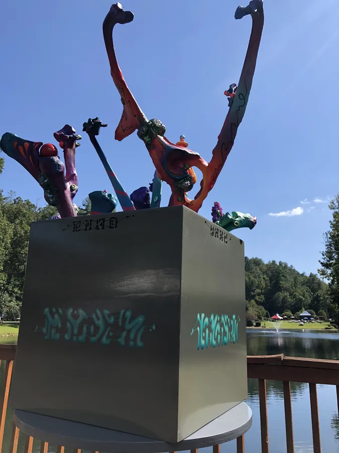 Magic Box from below: a grey cube with teal markings on its sides, orange, purple and blue stalks arching up from the top against a blue sky.