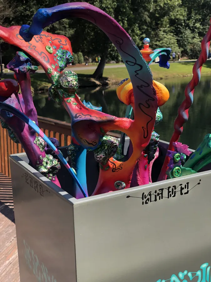 Detail of Magic Box: looking down into the open grey cube at tangled orange, magenta and blue stalks with small green creatures, the pond behind.