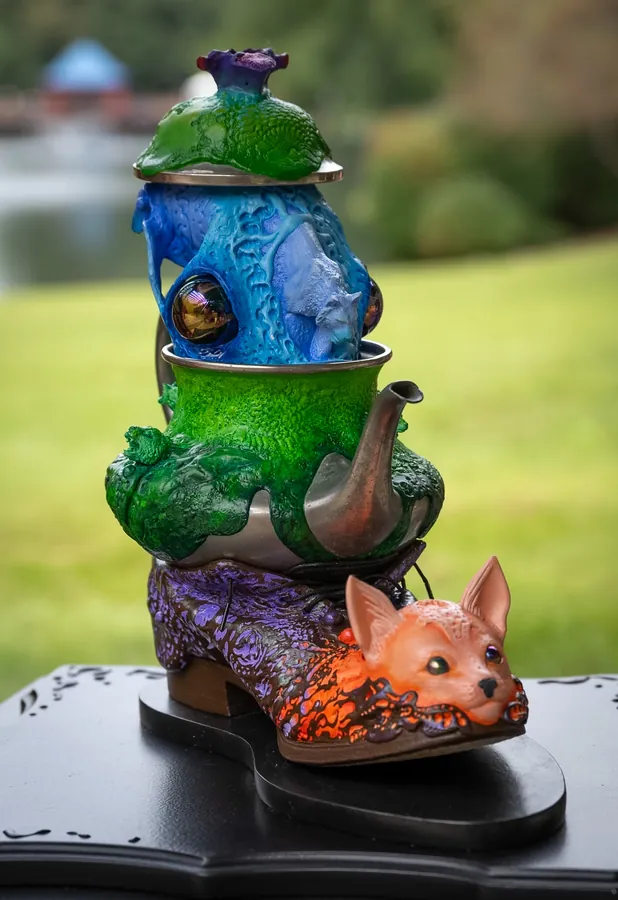 Detail of Pussyfoot: a purple and orange shoe with a fox face at the toe, under a green teapot, a blue textured vessel with a glass eye, and a green lid.