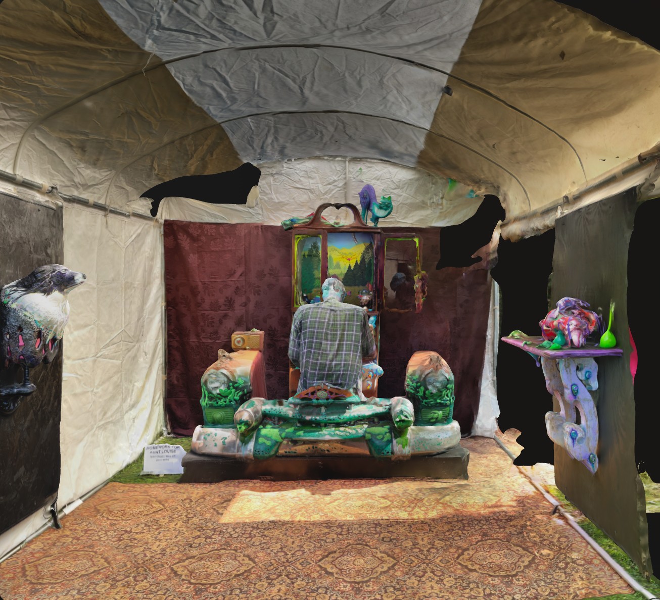 A 3D scan of the whole tent at the 2021 show: Homework for Aunt Louise, a pair of painted car fenders and a carved cabinet with a landscape inside, with Nat seated at it from behind, a wall-mounted animal head to the left and a painted shelf to the right.