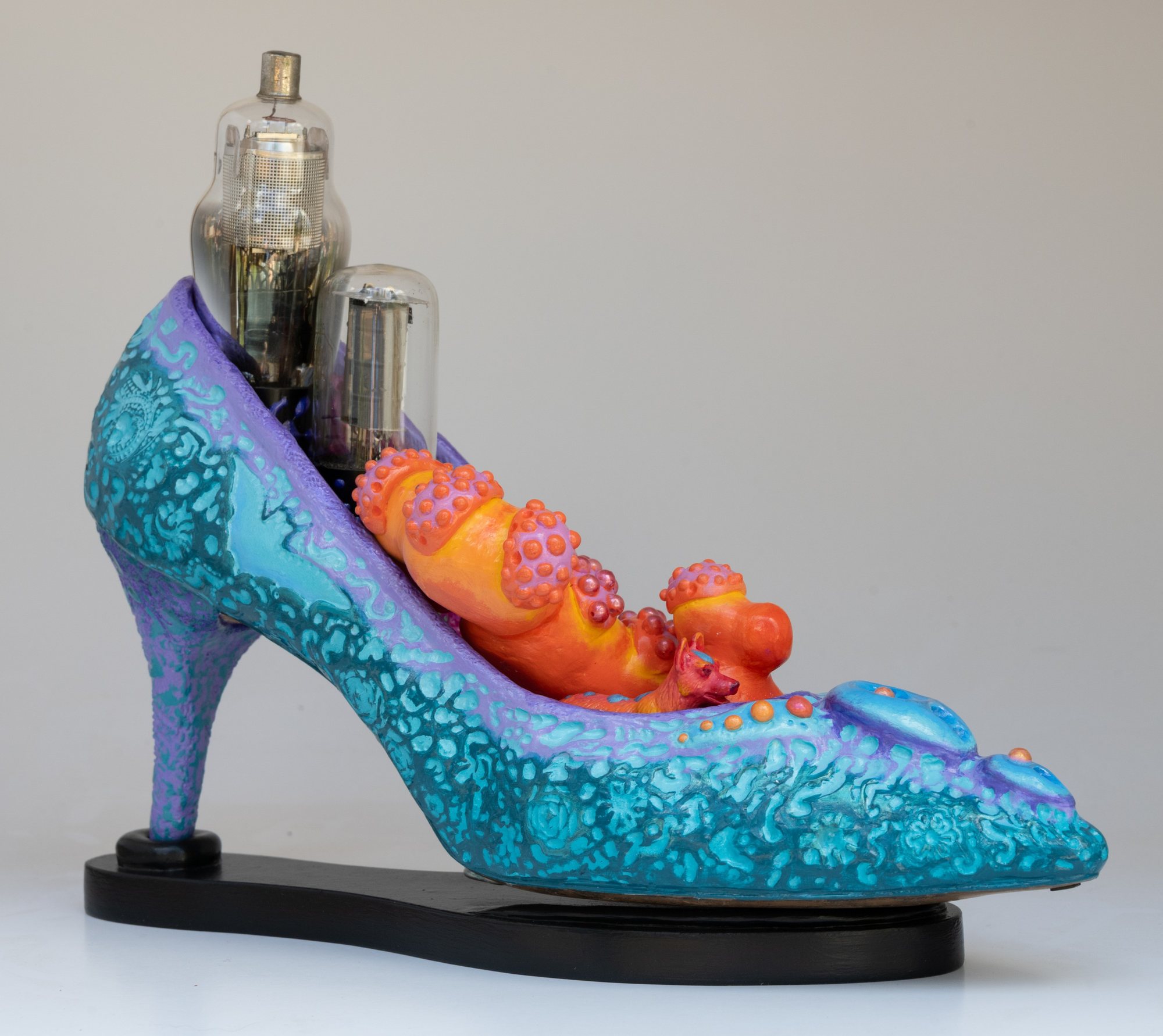 A turquoise high-heeled shoe sculpture with orange creatures nestled inside and vacuum tubes rising from the heel.
