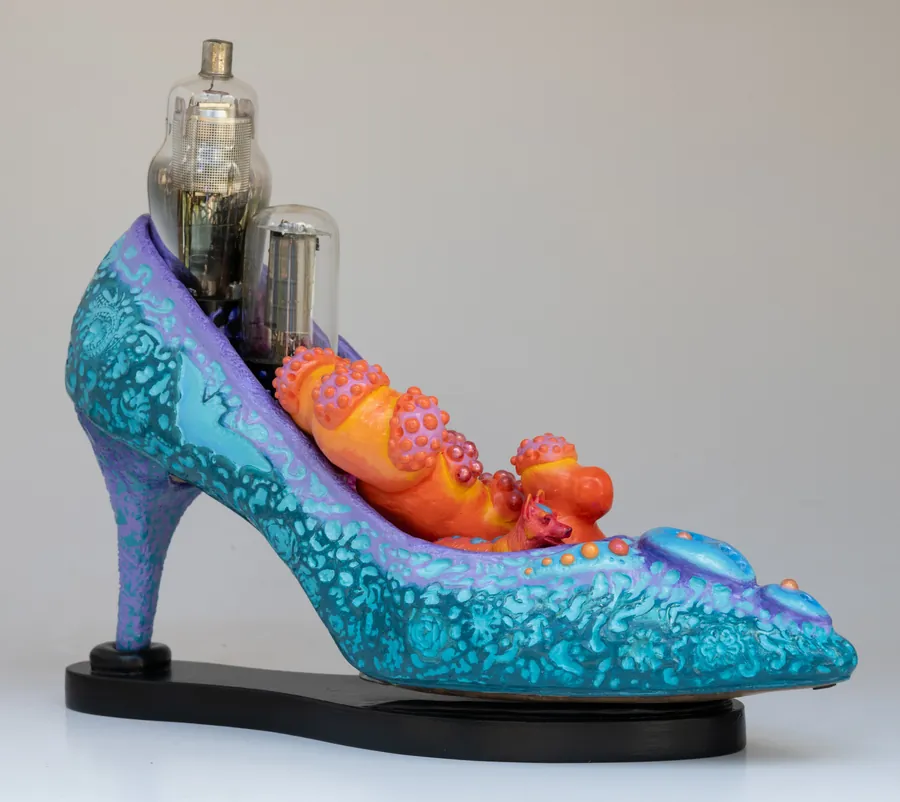 A turquoise high-heeled shoe sculpture with orange creatures nestled inside and vacuum tubes rising from the heel.