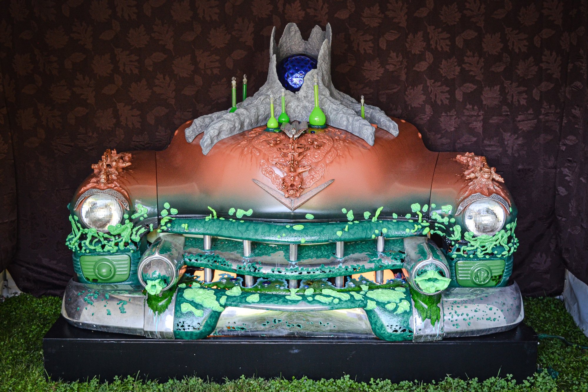 The front of a classic car painted green and coral, its grille dripping with green, topped by a carved stump with a blue eye.