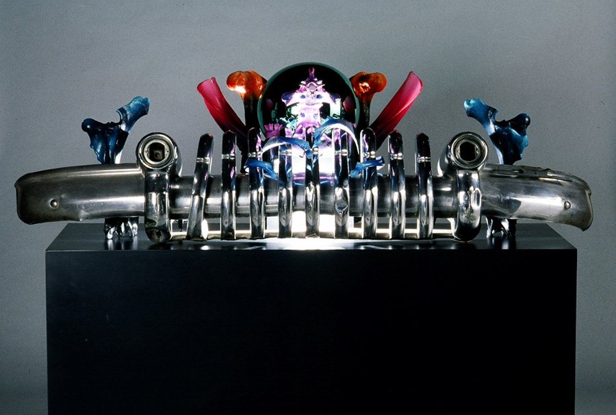 A chrome car bumper and grille on a black stand, topped with small colorful creatures.