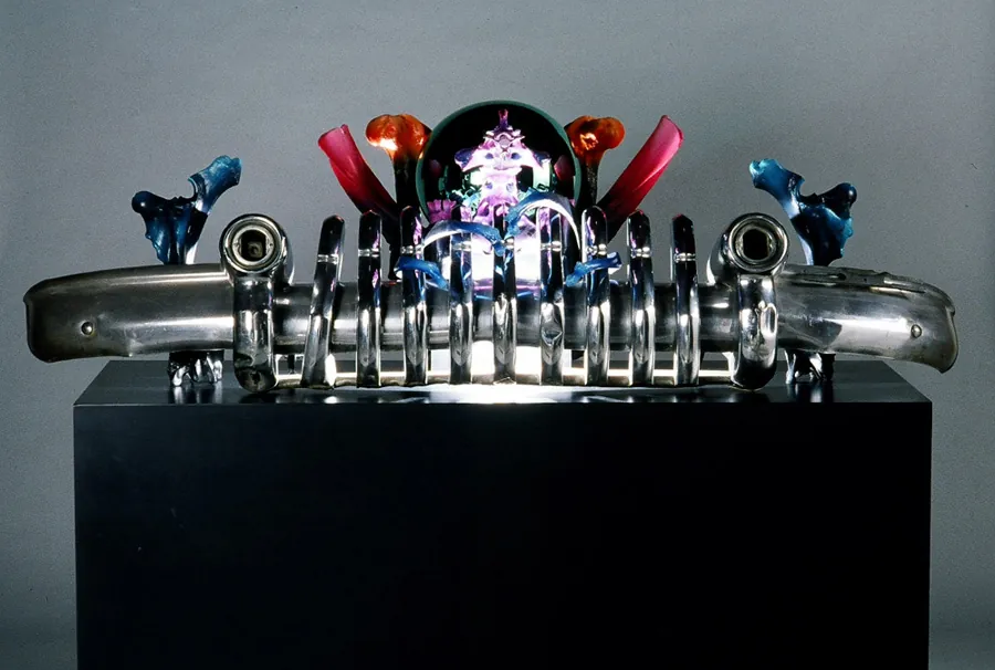 A chrome car bumper and grille on a black stand, topped with small colorful creatures.