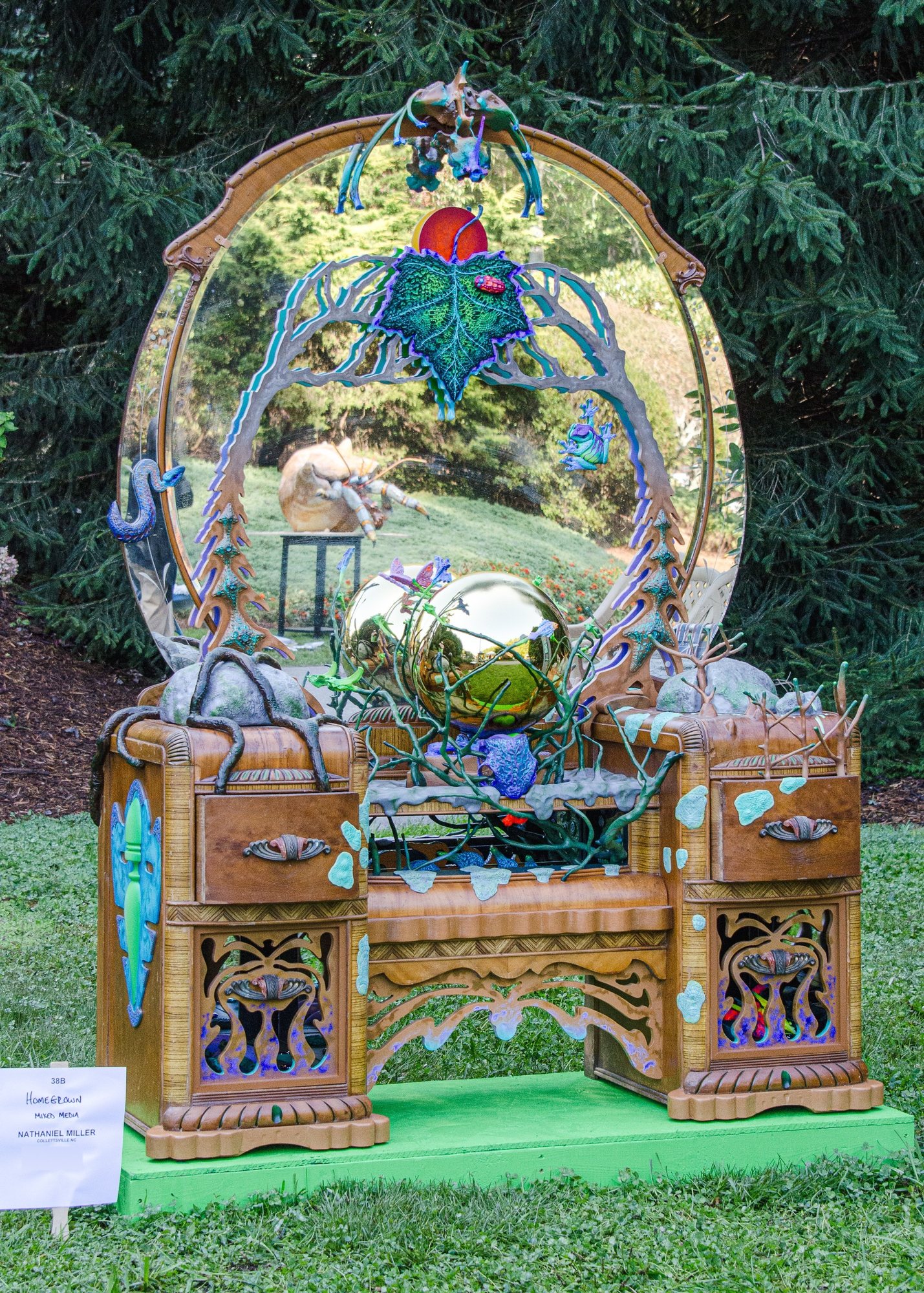 A carved wooden vanity with an oval mirror, wrapped in painted vines and leaves, holding a silver gazing ball.