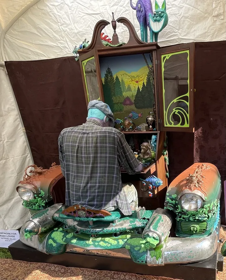 A figure with blue-grey skin, a flat cap and a plaid shirt sits at the open desk of a tall wooden hutch on the green, copper and chrome front end of an old car, a painted landscape glowing inside the hutch.
