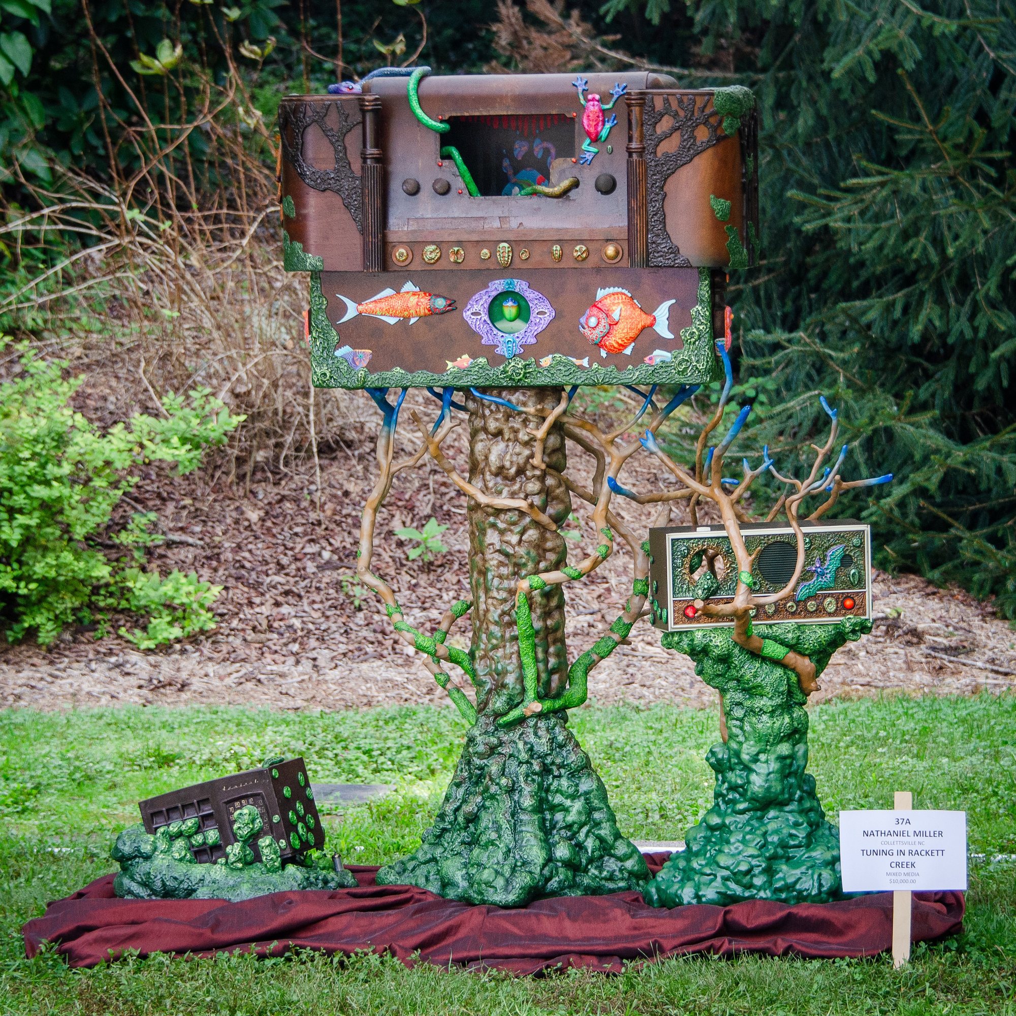 An outdoor sculpture: a painted wooden box with fish and a small stage stands on green, root-like trunks, beside a second small box.