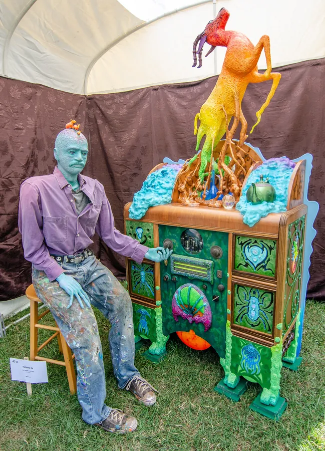 A blue-skinned figure in paint-spattered jeans sits on a stool at a carved green cabinet crowned by a rearing orange horse.